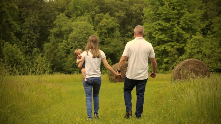 John Covington and his wife Kim walk hand in hand through a lush, grassy field toward round hay bales and a line of trees, with Kim carrying their young daughter on her hip.